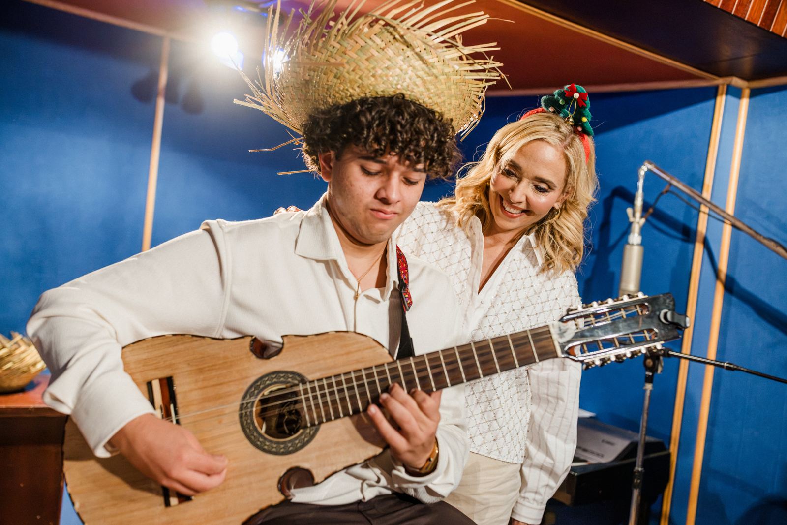 Un joven músico toca el cuatro puertorriqueño con sombrero de paja mientras Marisoll sonríe a su lado